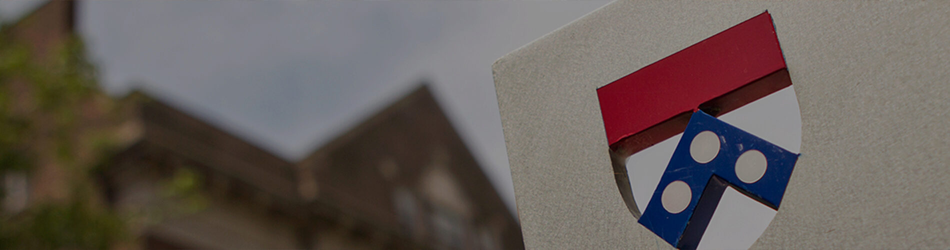 A close-up of a shield emblem with a red top, a blue section with white circles, and an abstract background of blurred architecture and foliage.