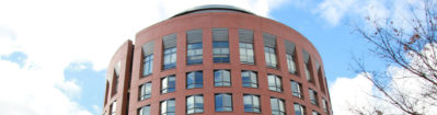 A round, modern building with red brick exterior and large windows, set against a partly cloudy sky, with tree branches visible on the right.