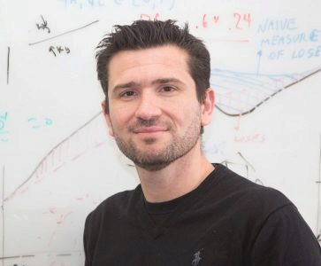 A person with short dark hair and a beard, wearing a black shirt, standing in front of a whiteboard with various graphs and notes.