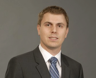 Headshot of a person wearing a dark suit, white shirt, and blue striped tie against a neutral gray background.