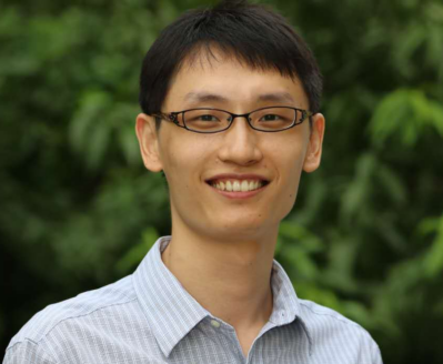 A headshot of a smiling person wearing glasses and a striped shirt, with a blurred green foliage background.