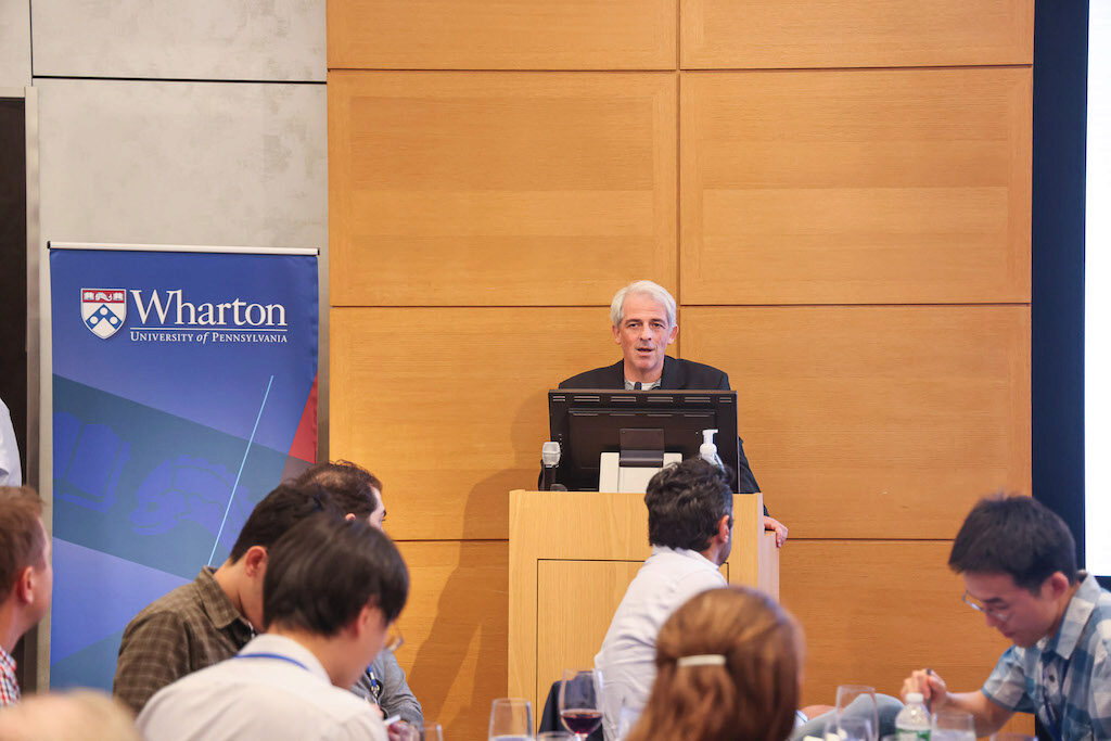 Itay Goldstein addresses an audience from a podium. A Wharton banner is to the viewer's left.