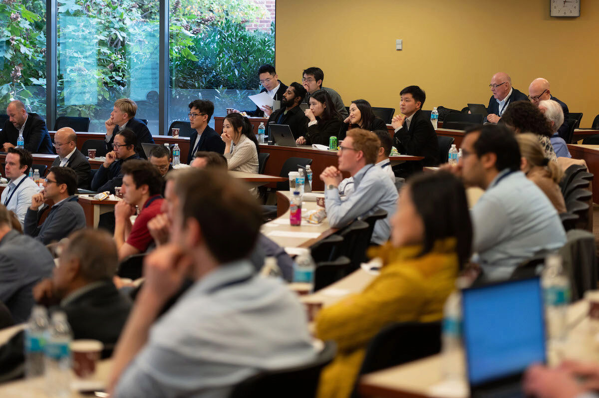 A group of people attentively listening during a seminar or lecture in a classroom setting. They are seated in tiered rows, some using laptops, and surrounded by notebooks and drinks.