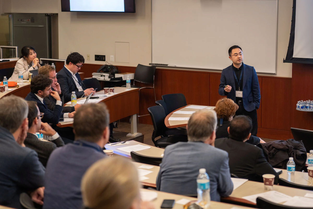 A person is giving a presentation in a lecture hall. An audience of several individuals is seated, some taking notes, others listening attentively. The setting is professional, with water bottles and papers on the tables.