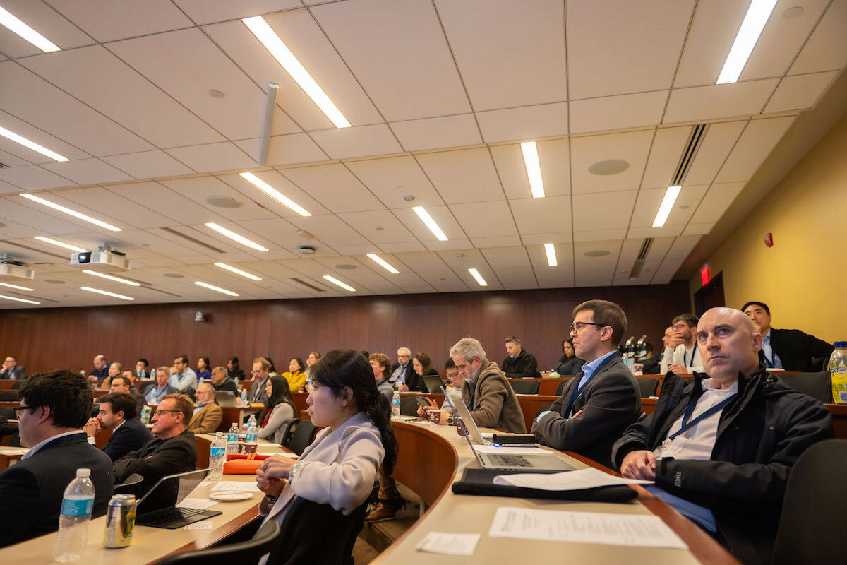 A group of people seated in a lecture hall, attentively listening to a presentation or lecture. The room is brightly lit with a wood-paneled wall and projection equipment visible.