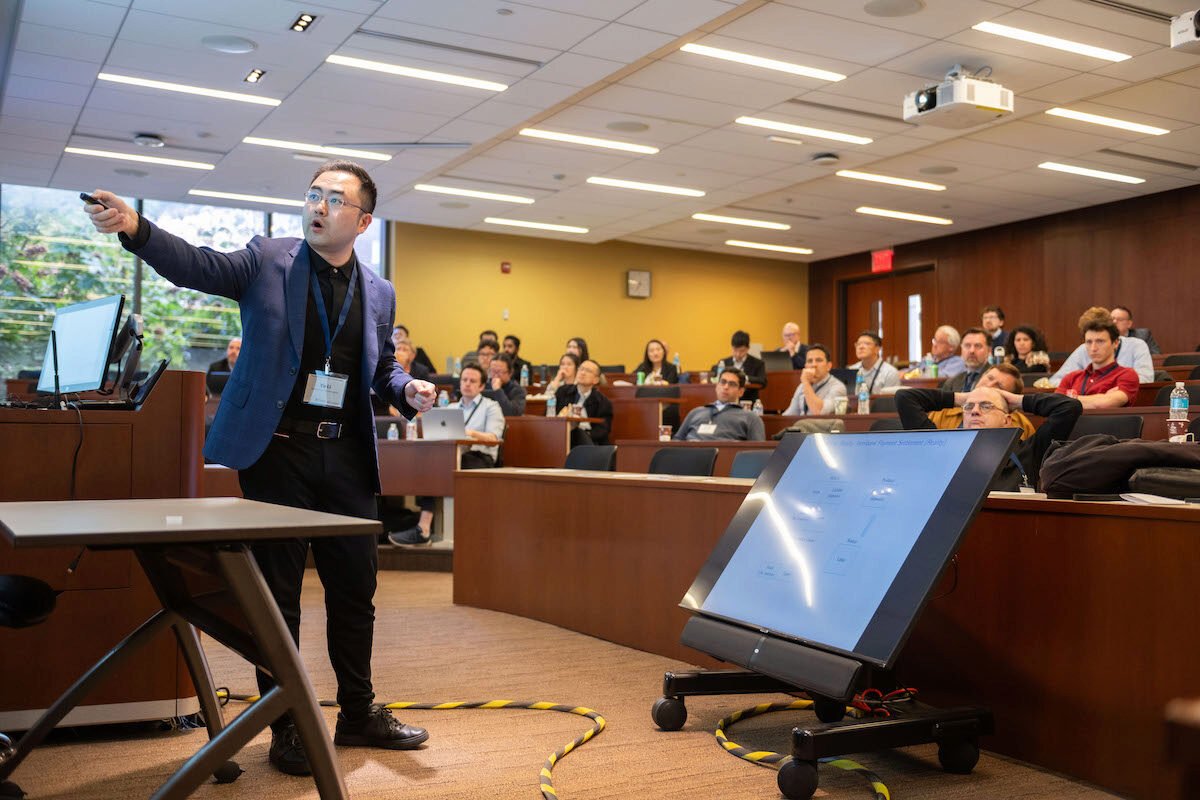 A person is giving a presentation in a lecture hall, pointing towards a screen. The audience, comprised of several people, is seated and attentively watching.