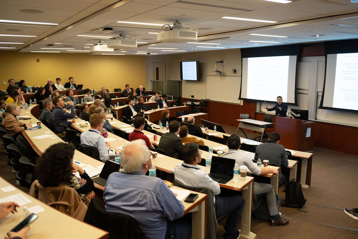 A lecture hall with a presenter speaking to an engaged audience. Attendees are seated in tiered rows, taking notes and using laptops.