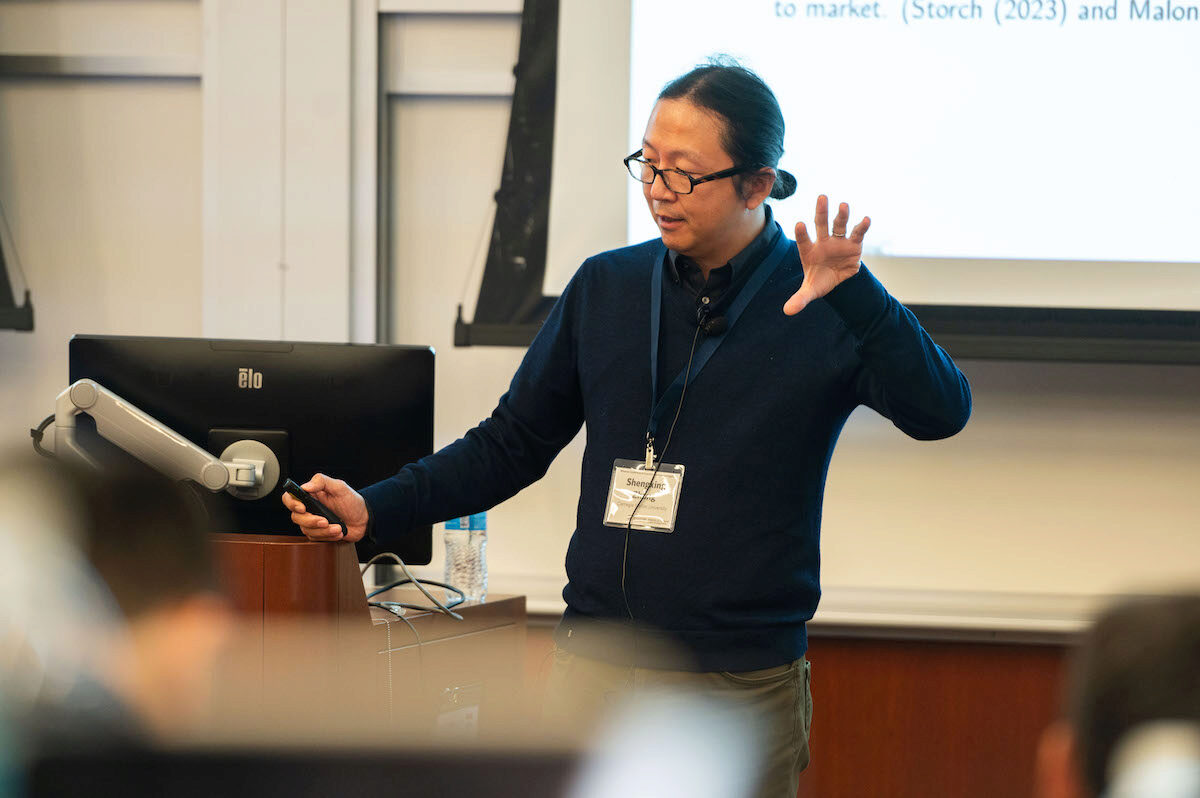 A person is giving a presentation in a classroom or lecture setting. They are standing near a computer monitor and gesturing with their hand, appearing to explain a concept.