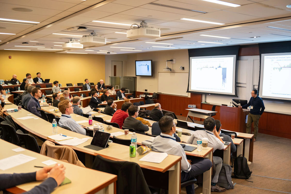 A classroom or lecture hall with several people seated at desks, attentively listening to a speaker presenting at the front. A projection screen displays a graph.