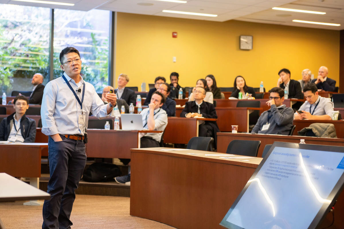 A person giving a presentation in a lecture hall while a small group of people listen attentively. A large screen displays presentation slides.