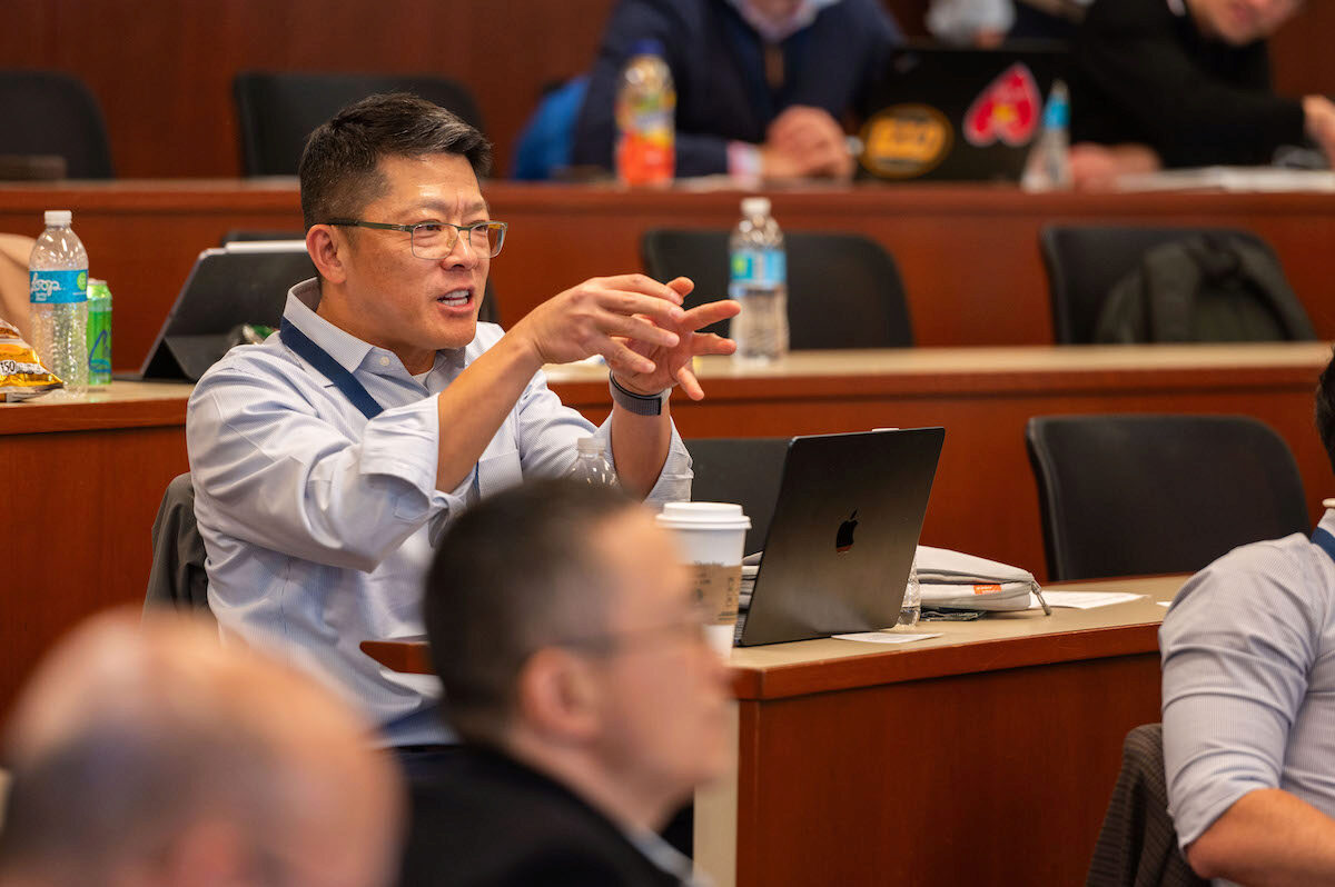 A person is seated in a classroom setting, engaged and gesturing while participating in a discussion. A laptop, water bottles, and a coffee cup are visible on the desk. Other individuals are in the background, focused on their own devices.