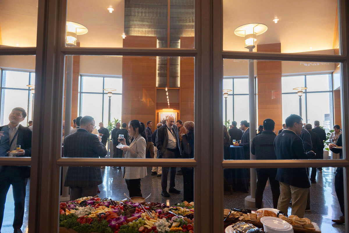 A gathering of people in a stylish indoor venue, featuring a large buffet table with assorted fruits and appetizers. The group appears to be mingling at a social or professional event.