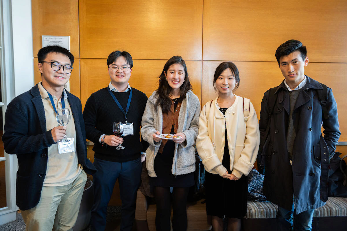 A small group of people smiling, standing together indoors. Some are holding glasses, suggesting a social gathering or event. They are dressed in smart casual attire.