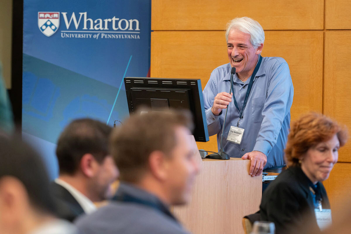 A person speaking at a podium with a microphone, smiling, in front of a Wharton School University of Pennsylvania banner. Several attendees are listening.