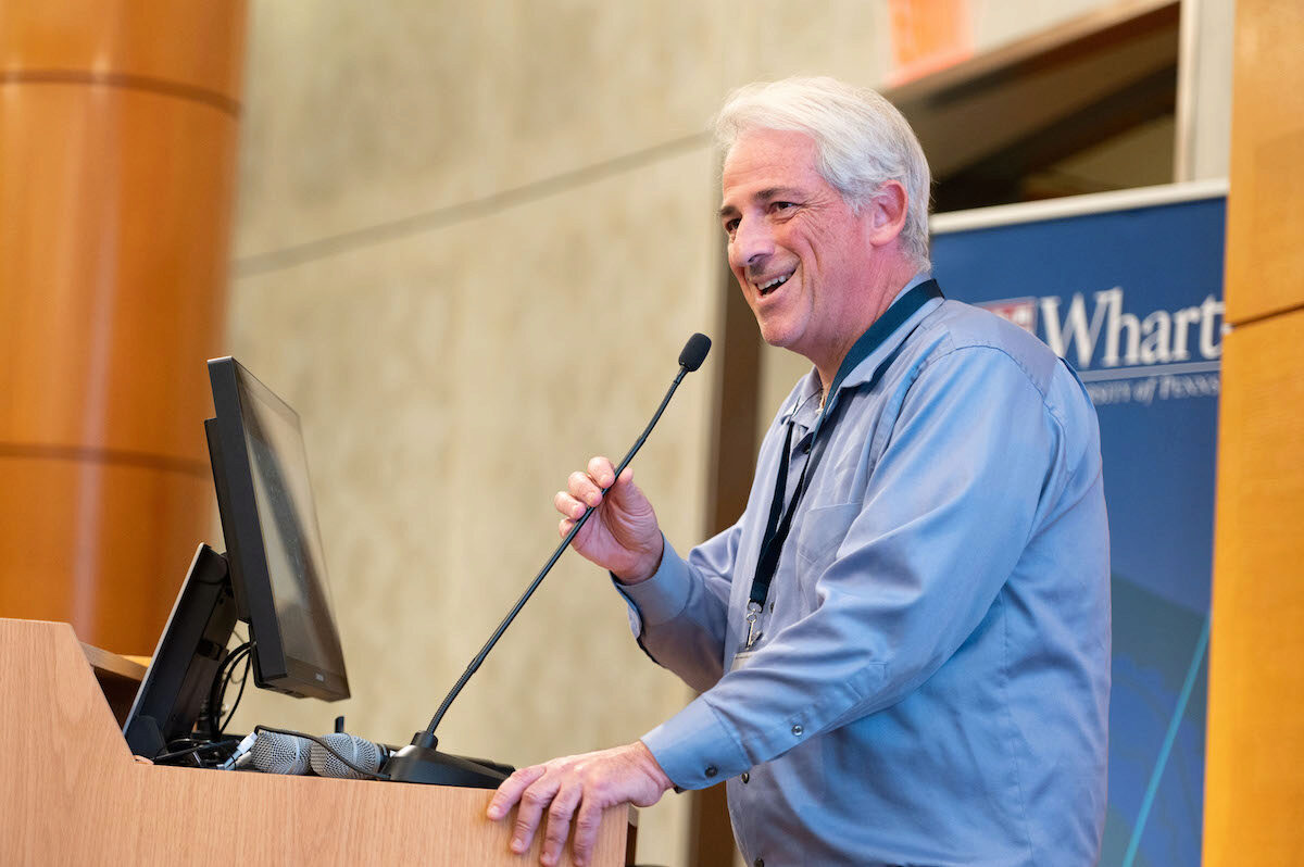 A person speaking at a podium with a microphone and a computer screen, set against a backdrop with the Wharton School logo.
