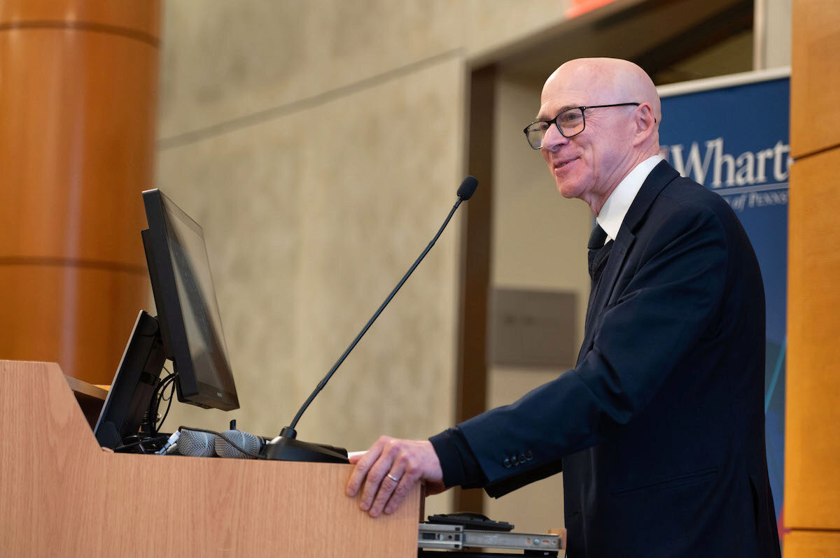 A person wearing glasses, speaking at a podium with a microphone, in a formal setting. A banner with "Wharton" is visible in the background.