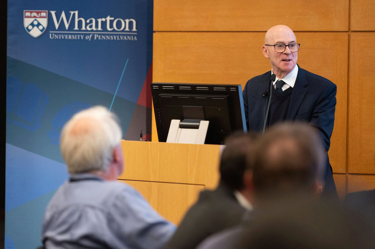 A person in a suit and glasses is speaking at a podium with a microphone. A banner for the Wharton School, University of Pennsylvania, is in the background. Audience members are blurred in the foreground.