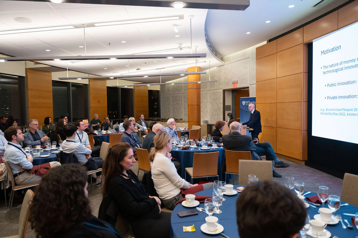 A speaker is giving a presentation to a seated audience in a conference room. Attendees are seated at round tables with blue tablecloths, and a large screen displays the presentation topic.