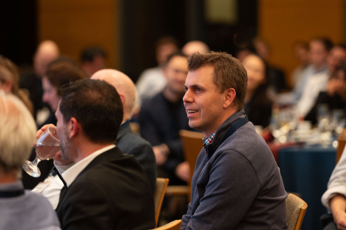 A person in a blue sweater listens attentively at a conference or seminar. The background shows other attendees and tables with glasses and plates.