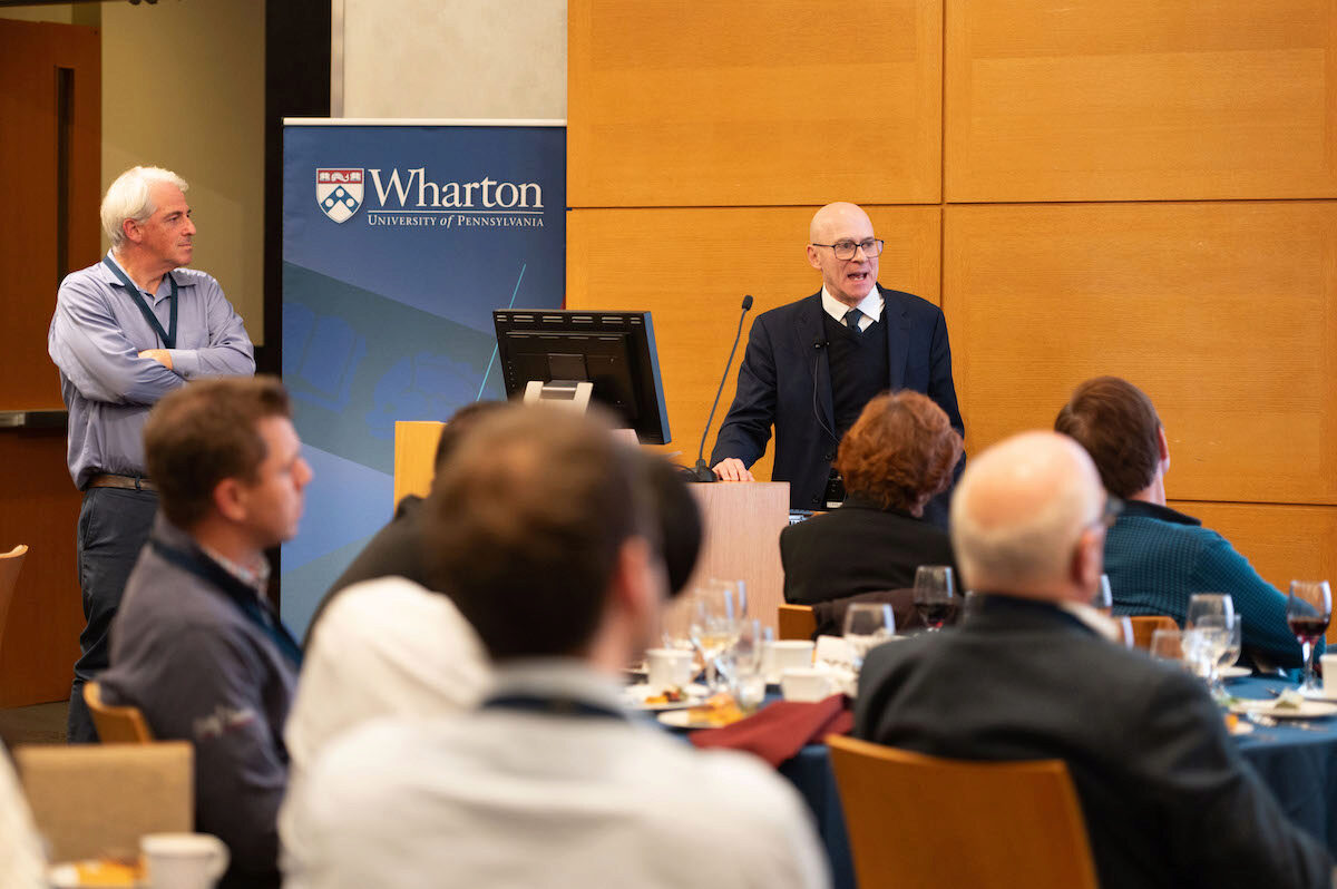 A person in a suit speaks at a podium in a conference room, with an audience seated at tables. A Wharton University of Pennsylvania banner is visible.