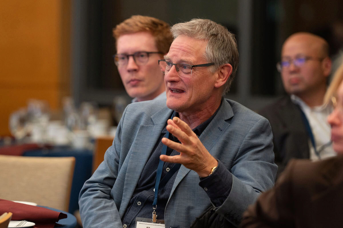 A discussion at a conference or meeting, with a person in the foreground speaking and gesturing, and others attentively listening in the background.