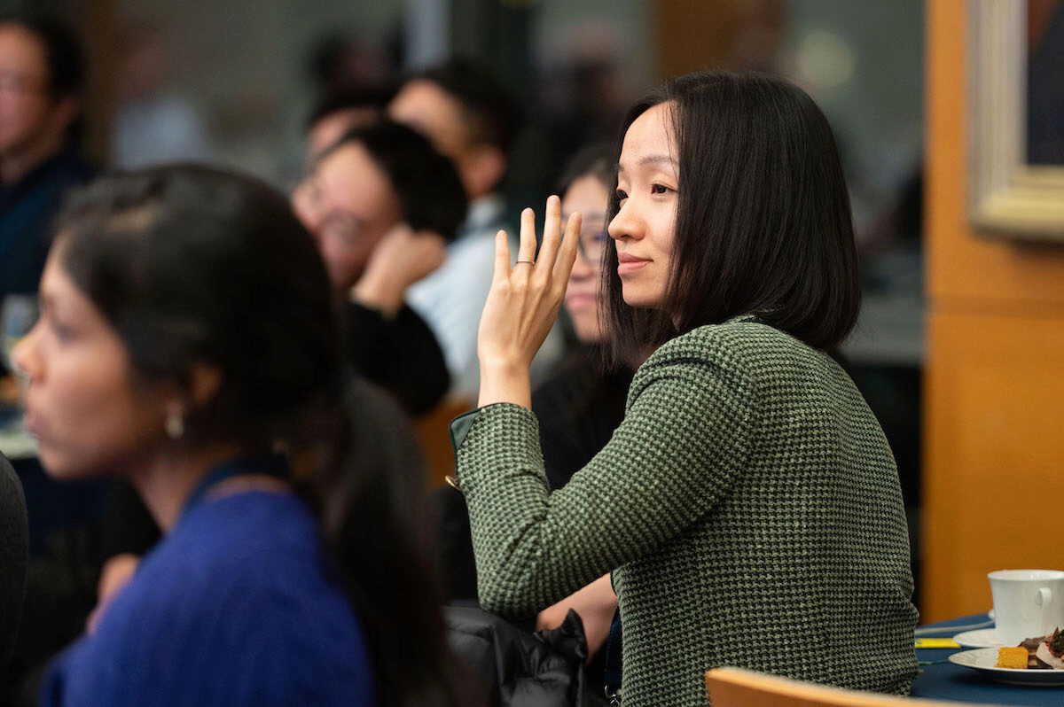 A small group of people seated at a gathering or event, with one person raising a hand as if asking a question or participating. The environment suggests a seminar or conference setting.