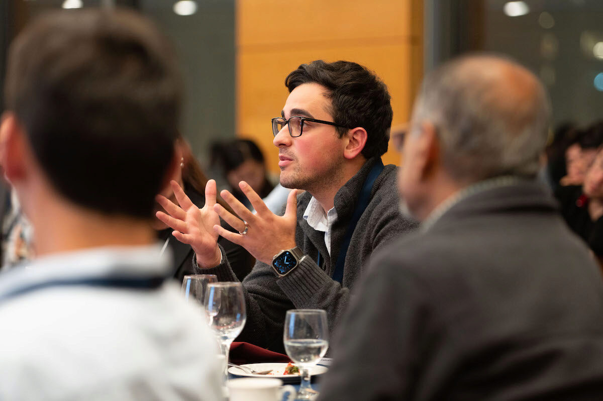 A person gesturing while speaking at a meeting or conference. They are seated at a table with glasses and cups, surrounded by other attendees.
