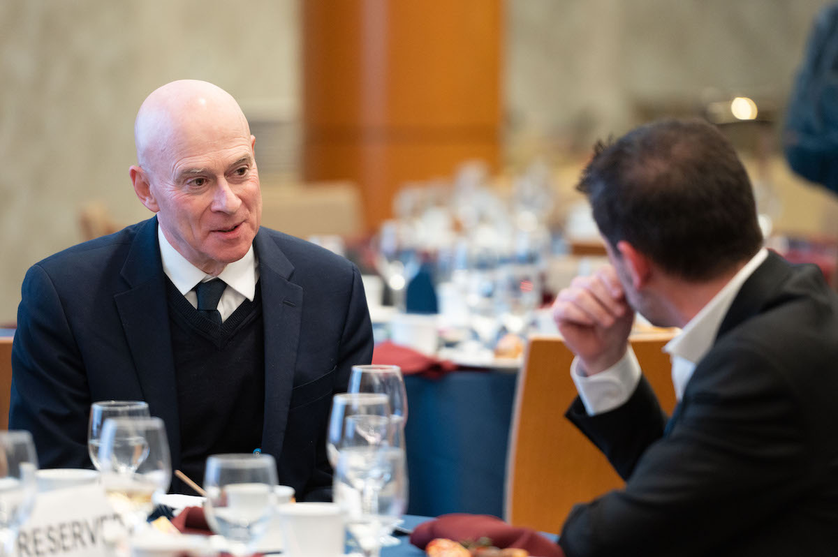 Two individuals in formal attire are engaged in conversation at a dining table set with glasses and plates. The background suggests a formal event or meeting.