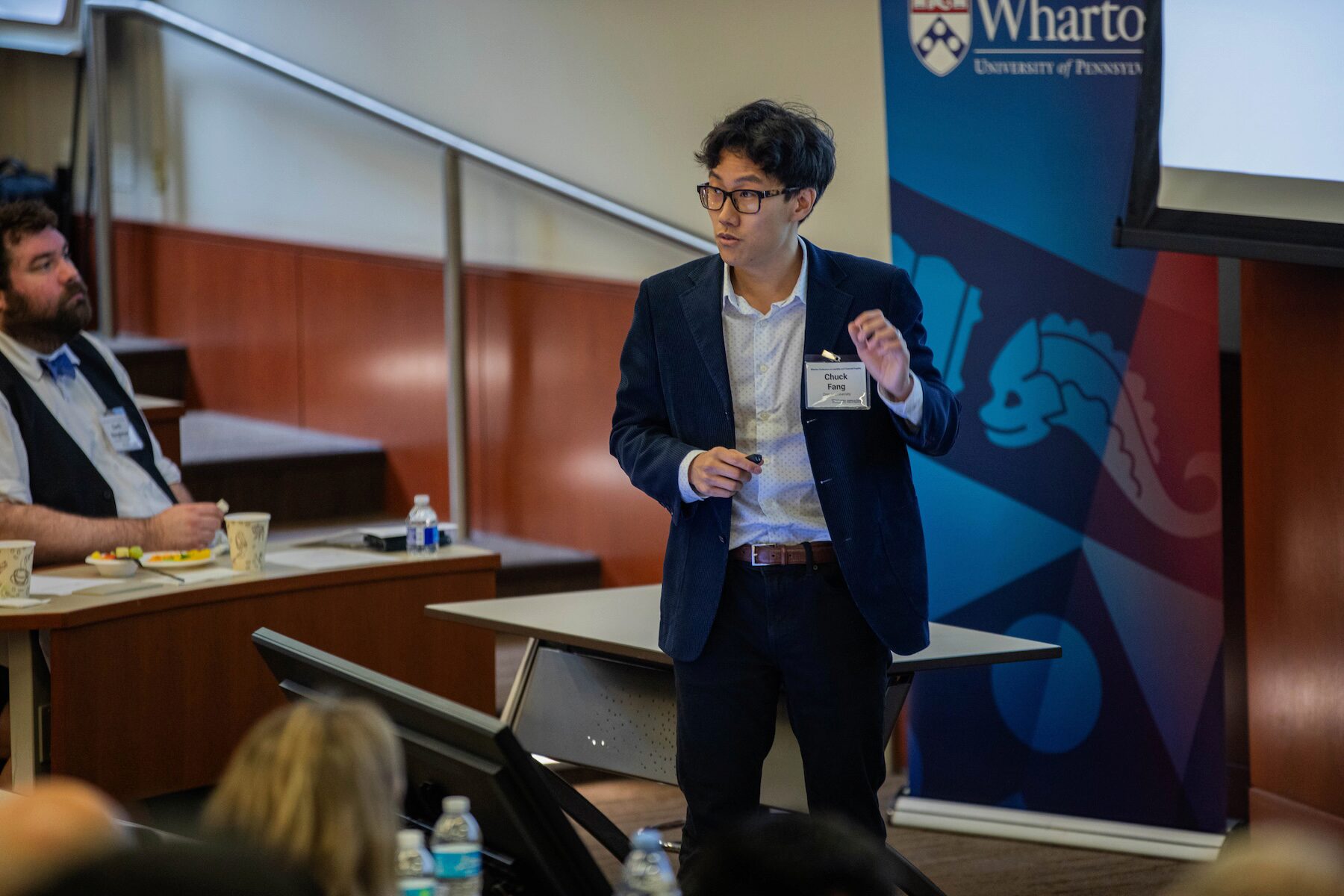 A speaker addresses an audience in a classroom before a Wharton blue banner