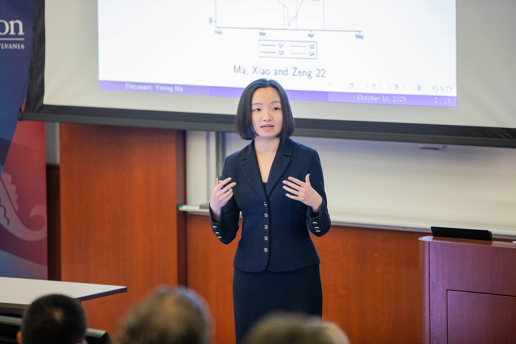 A speaker addresses an audience before a white screen in a classroom